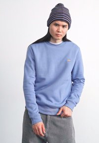 Light blue sweatshirt with a crew neck and small logo on the chest, paired with a multicolored knitted beanie. Textured gray corduroy pants.