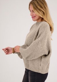 Lichtgrijze oversized trui met gestructureerde breisel, ingezette schoudernaden en geribbelde manchetten. Gedragen over een donkere spijkerbroek met meerdere armbanden.