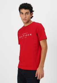 Raudona medvilninė marškinė su trumpomis rankovėmis, su baltu "TOMMY HILFIGER" logotipu. Lygi tekstūra, apvalus iškirpimas ir mažas logotipo akcentas ant rankovės.