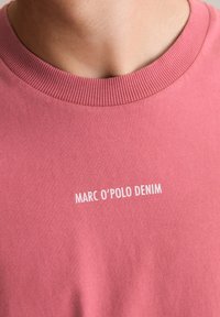 Roze katoenen T-shirt met ronde hals, met een witte logo-opdruk die "MARC O'POLO DENIM" op de voorkant leest. Zachte textuur, casual ontwerp.