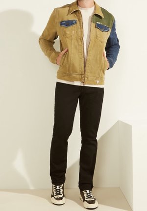 Denim spijkerjack met een beige lijf, blauwe en groene schouderpanelen, twee borstzakken, ritssluiting, gedragen over een gestructureerde witte trui en zwarte broek.