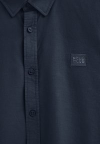 Camicia corta in navy realizzata in tessuto testurizzato, con una chiusura a bottoni e un'etichetta quadrata con il logo "POLO CLUB" in un navy più scuro.