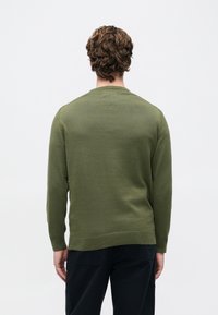 Άνδρας με σγουρά καστανά μαλλιά, φορεμένος σε ελιά πράσινο πουλόβερ και μαύρο παντελόνι, στέκεται μπροστά από ένα απλό λευκό φόντο.