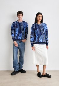 Des pulls en tricot bleu avec des motifs abstraits, associés à des jeans clairs et une jupe blanche. Les modèles portent des chaussures noires, soulignant le contraste et la texture.