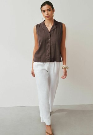 Vrouw die een bruine mouwloze blouse met knopen draagt, witte wijd uitlopende broek, beige sandalen en gouden oorbellen, staand tegen een eenvoudige lichte muur.