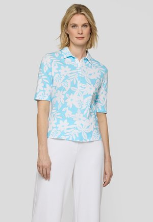 Frau trägt ein hellblaues Poloshirt mit kurzen Ärmeln und weißen Blumenmustern sowie weiße Hosen mit weitem Bein vor einem einfarbigen Hintergrund.