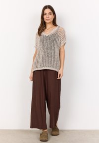 Knit beige top with a loose fit and V-neck, paired with brown wide-leg pants and green slip-on shoes, on a model against a plain background.