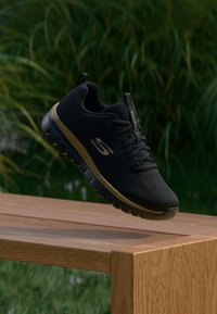 Schwarzer Sportschuh mit einem strukturierten Obermaterial aus Stoff, dünnen Schnürsenkeln und einer gepolsterten Sohle. Verfügt über ein dezentes Logo und eine goldfarbige Akzent-Mittelsohle.