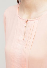 Blusa rosa chiaro realizzata in tessuto morbido, con scollatura aderente, dettaglio dei bottoni e pieghe verticali sulla parte anteriore per un tocco di textura.