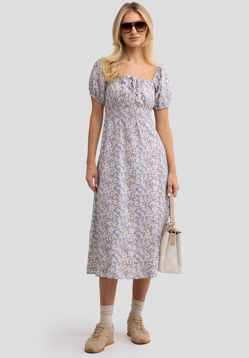 Vestido floral con mangas abullonadas, escote cuadrado, patrón en azul claro y beige. La modelo sostiene un bolso crema, llevando sneakers y calcetines color arena.