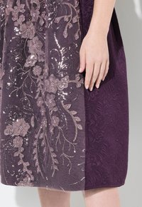 Paarse jurk met geborduurde bloemenpatronen met pailletten en een getextureerd lijfje. Eén zijde is doorschijnend, waardoor de ontwerpdetails extra opvallen.