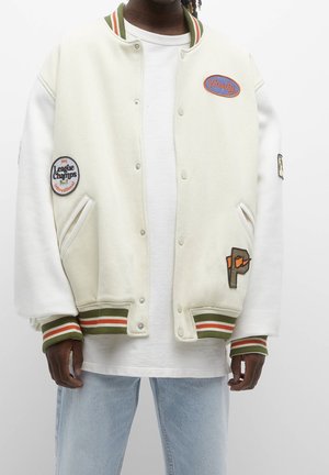 Person trägt eine cremefarbene Varsity-Jacke mit grün und orange gestreiften Bündchen sowie Aufnähern mit den Schriftzügen „Varsity“ und „League Champs“ über einem weißen Hemd.