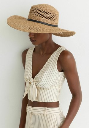 Nainen, jolla on päällään beige leveälierinen punottu hattu ja ylleen sopiva kermaisen värinen lyhyt paita ja housut, joissa on ohuet mustat pystysuorat raidat.