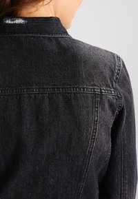 Giacca di jeans nera con colletto, yoke posteriore e cuciture a vista, che presenta un tessuto leggermente texturizzato con una finitura morbida.