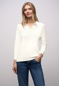 Maglia bianca con zip, maniche lunghe e collo con risvolto, abbinata a jeans blu scuro. Texture liscia e design casual.
