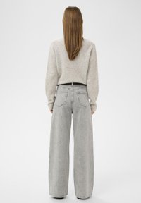 Žena s rovnými hnědými vlasy, oblečená v světle šedém krátkém svetru a vyměřených širokých džínách s vysokým pasem, stojí zády.