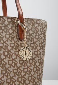 Sac fourre-tout beige avec un motif de logo répétitif, poignées en cuir marron et une étiquette en métal doré attachée par une chaîne. Extérieur en tissu texturé.