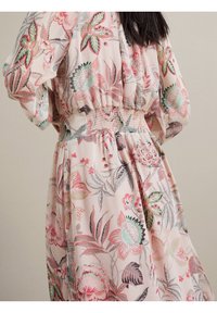 Femme portant une robe florale rose avec une taille smockée et des manches bouffantes, debout devant un fond beige uni.