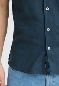 Chemise en lin bleu marine avec un devant boutonné, des boutons blancs et une étiquette sur le côté, offrant une coupe décontractée et une texture lisse.