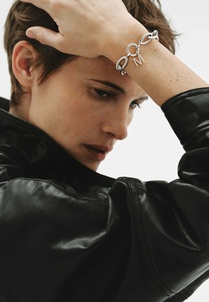 Primer plano de una persona con chaqueta de cuero negra, apoyando su mano en la frente, mostrando una pulsera de cadena plateada con un colgante en forma de 'M'.