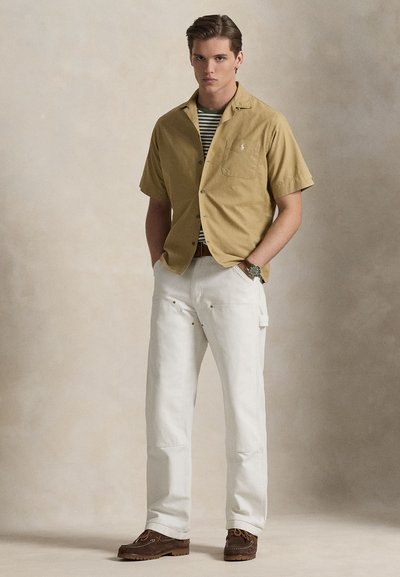 Μπλούζα με κοντά μανίκια και κουμπιά, ριγέ μπλουζάκι από κάτω, λευκό παντελόνι φαρδιάς γραμμής και καφέ παπούτσια τύπου slip-on, όρθιος σε ουδέτερο φόντο.