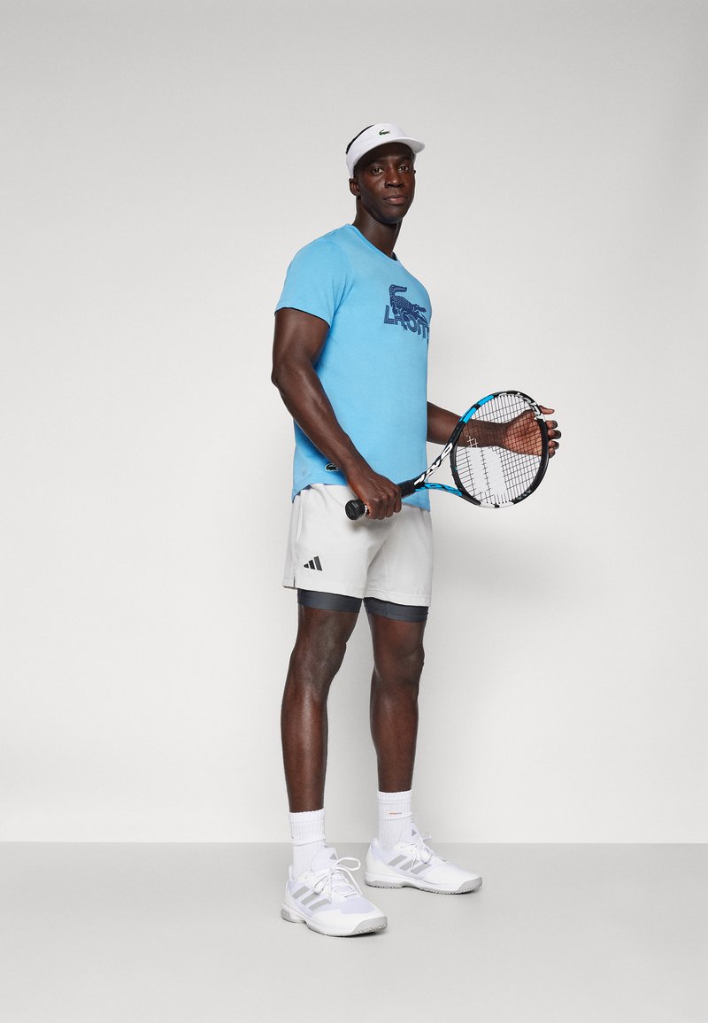 Blauwe t-shirt met korte mouwen met een donkerblauw logo, lichtgrijze shorts met een zwarte tailleband, witte tennisschoenen en een tennisracket in de hand.