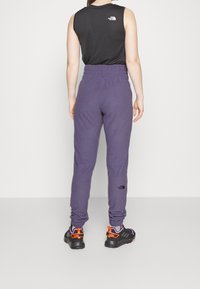Schwarzes ärmelloses Oberteil, lila Sweatpants mit elastischem Bund und schwarze Sportschuhe mit orangefarbenen Akzenten, sichtbaren Markenlogos.