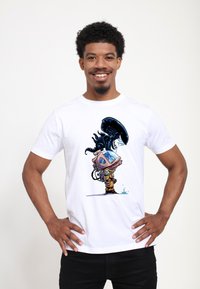 Muž s úsměvem, rukama v bok, v bílé košili, ukazuje kreslenou mimozemskou bytost přilnutou k astronautovi v helmě stojícímu u kapky vody.