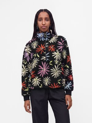 Femme avec de longues tresses portant un pull polaire noir avec des motifs floraux colorés et un col à fermeture éclair mi-hauteur, associé à un pantalon noir.