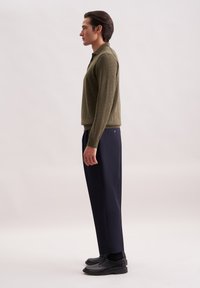 Maglione verde oliva, vestibilità slim, polsini a coste; abbinato con pantaloni blu scuro, taglio dritto, caratterizzati da un dettaglio con bottone sulla tasca posteriore, scarpe nere.