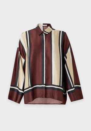 Femme debout les mains dans les poches, portant une chemise à rayures oversize, un pantalon blanc, une ceinture marron, des chaussures vertes et des boucles d'oreilles créoles dorées sur fond blanc.