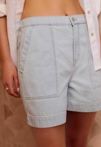 Shorts en denim bleu clair à taille haute, dotés de poches latérales, de bords roulés et d'une fermeture à bouton sur le devant. Texture douce et design épuré.