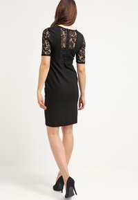 Svart klänning med korta ärmar i blommönstrad spets, en figurnära silhuett med midilängd och en subtil detalj på bakåtslutningen.