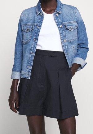 Person iført en lyseblå denimjakke med opkrogede ærmer, hvid t-shirt med rund hals og højtsiddende, sorte skræddersyede shorts.