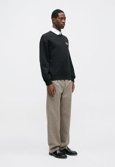 Čierna mikina s bielym logom, šedé textúrované nohavice a čierne topánky. Model stojí v profile na pozadí bez vzoru.