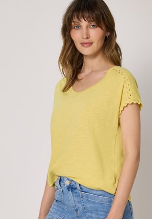Femme aux cheveux bruns mi-longs, portant un t-shirt jaune à manches courtes avec des bords festonnés et un jean bleu clair, devant un fonds uni.