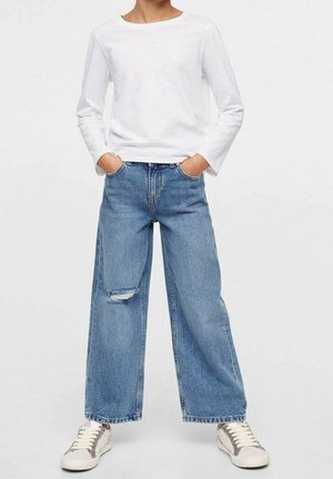 Personne portant un haut blanc à manches longues, un jean bleu ample avec une déchirure au genou gauche, et des baskets blanches avec des rayures beiges.