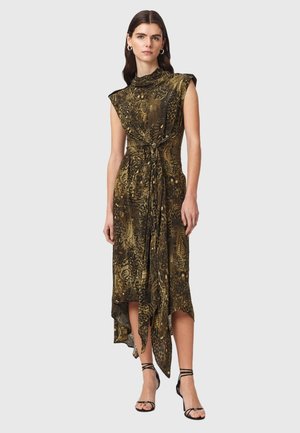 Femme portant une robe midi sans manches imprimé animalier vert olive, ourlet asymétrique, taille nouée et talons noirs à brides, debout devant un fond gris.
