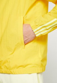Žlutá lehká bunda s texturovaným povrchem, s boční kapsou a třemi vertikálními bílými pruhy na rukávu.