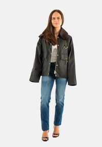 Dunkelgrüne wasserdichte Jacke mit einem braunen Cordkragen, Fronttaschen und goldenen Beschlägen, kombiniert mit blauen Jeans und hohen Schuhen.