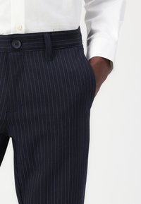 Man in donkere marineblauwe pinstripe broek en witte overhemd met hand in broekzak tegen een vlakke achtergrond.