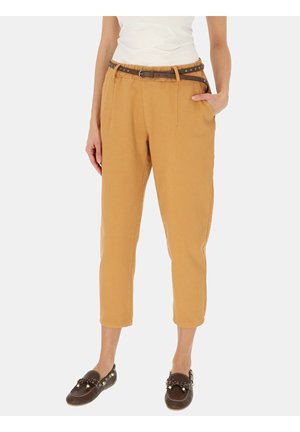 Kobieta w przyciętych musztardowych spodniach, brązowym pasku z ćwiekami, białym bezrękawnym topie i ciemnobrązowych mokasynach z frędzlami. Jedna ręka w kieszeni.