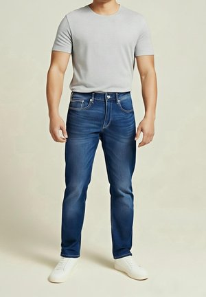 Man draagt een lichtgrijs T-shirt met korte mouwen, blauwe slim-fit jeans en witte sneakers, staand tegen een effen beige achtergrond.