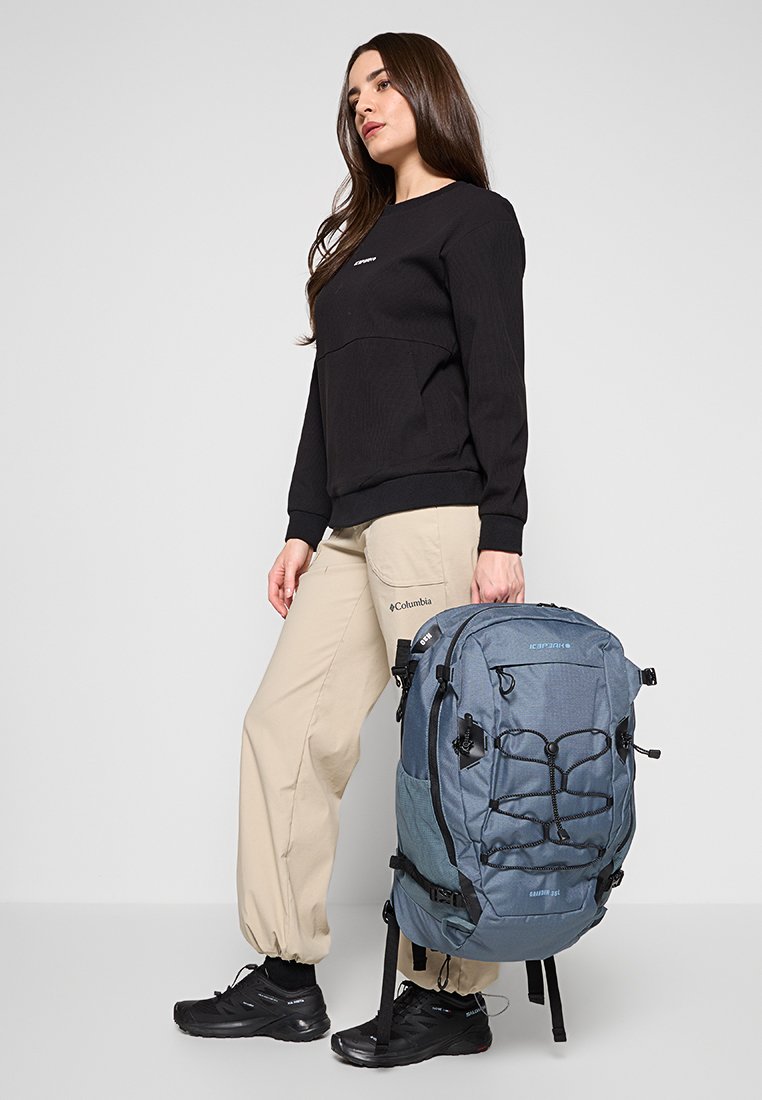 Blauer Wanderrucksack mit strukturierter Oberfläche, verstellbaren Trägern und Bungee-Kordel auf der Vorderseite. Modell trägt einen schwarzen Pullover und beige Hosen.