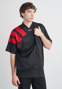 Čierne športové tričko s červenými pruhmi, krátkymi rukávmi a výstrihom do V. Modelka nesie čiernu kabelku cez rameno s vyrazeným logom.