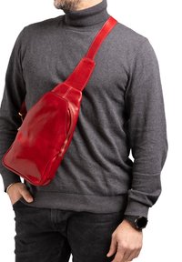 Borsa a tracolla rossa in pelle con finitura liscia, tracolla regolabile e tasca laterale con zip, indossata su un pullover grigio con collo alto.