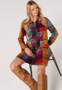 Žena sedící na židli, oblečená v barevných šatech na zip se vzorem patchwork a hnědých kozačkách po kolena, která se mírně dívá na stranu.