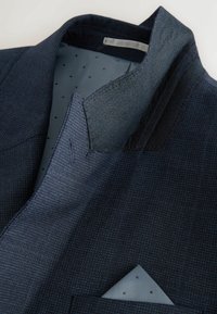 Navy blazer van een textuurstof, met een contrasterende kraag van grijze vilt, en een lichtblauwe voering met kleine zwarte stippen en een pochet.