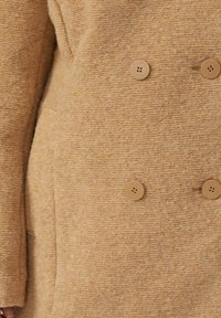 Doppelt-knöpfiger Mantel aus hellbeigem Stoff mit strukturierter Oberfläche. Verfügt über große runde beige Knöpfe und Seiten Taschen.