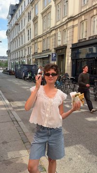 Young woman wearing sunglasses, white lace top, and denim shorts holds wrapped food, standing on a European city street with bikes and buildings.